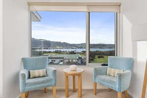 A room with two armchairs, a small round table, and a large window offering a view of a landscape with hills and a body of water. - Apollo Blue 12 (Apollo Bay)