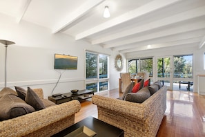 A spacious living room with high ceilings, large windows, wicker furniture, and a flat-screen TV mounted on the wall.