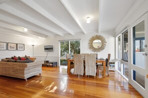 A spacious living room with a wooden floor, a sofa, a dining table with chairs, and a large window providing natural light. - Beach Fig (Skenes Creek, Apollo Bay)