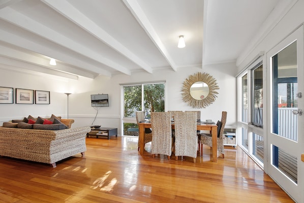 A spacious living room with a wooden floor, a sofa, a dining table with chairs, and a large window providing natural light. - Beach Fig (Skenes Creek, Apollo Bay)