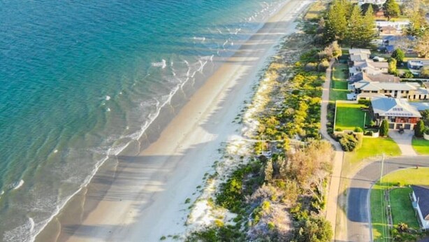 Eine Luftaufnahme eines Küstenwohngebiets mit Häusern direkt am Strand, einer klaren Straße und einem großen Gebäude mit markantem Dach.