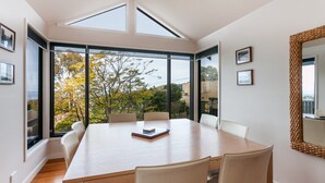 A modern dining room with a large table, chairs, and a view of trees through large windows. - Carrageen (Lorne)