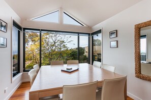 A modern dining room with a large table, chairs, and a view of trees through large windows. - Carrageen (Lorne)