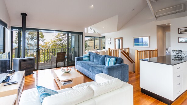 A spacious living area with a sofa, coffee table, and a fireplace. There is a balcony with a view of trees and a clear sky. - Carrageen (Lorne)