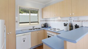 A modern kitchen with wooden cabinets, a built-in dishwasher, a stove, and a window with a view of greenery. - Beach Retreat (Apollo Bay)