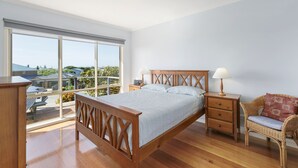 A bedroom with a large window, a wooden bed, a nightstand, a chair, and a ceiling light. - Beach Retreat (Apollo Bay)