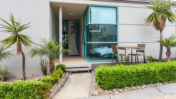 A modern building with a glass entrance, a small patio with a table and chairs, and a well-maintained garden with palm trees and shrubs. - Anglesea River Apartment 23 (Anglesea)