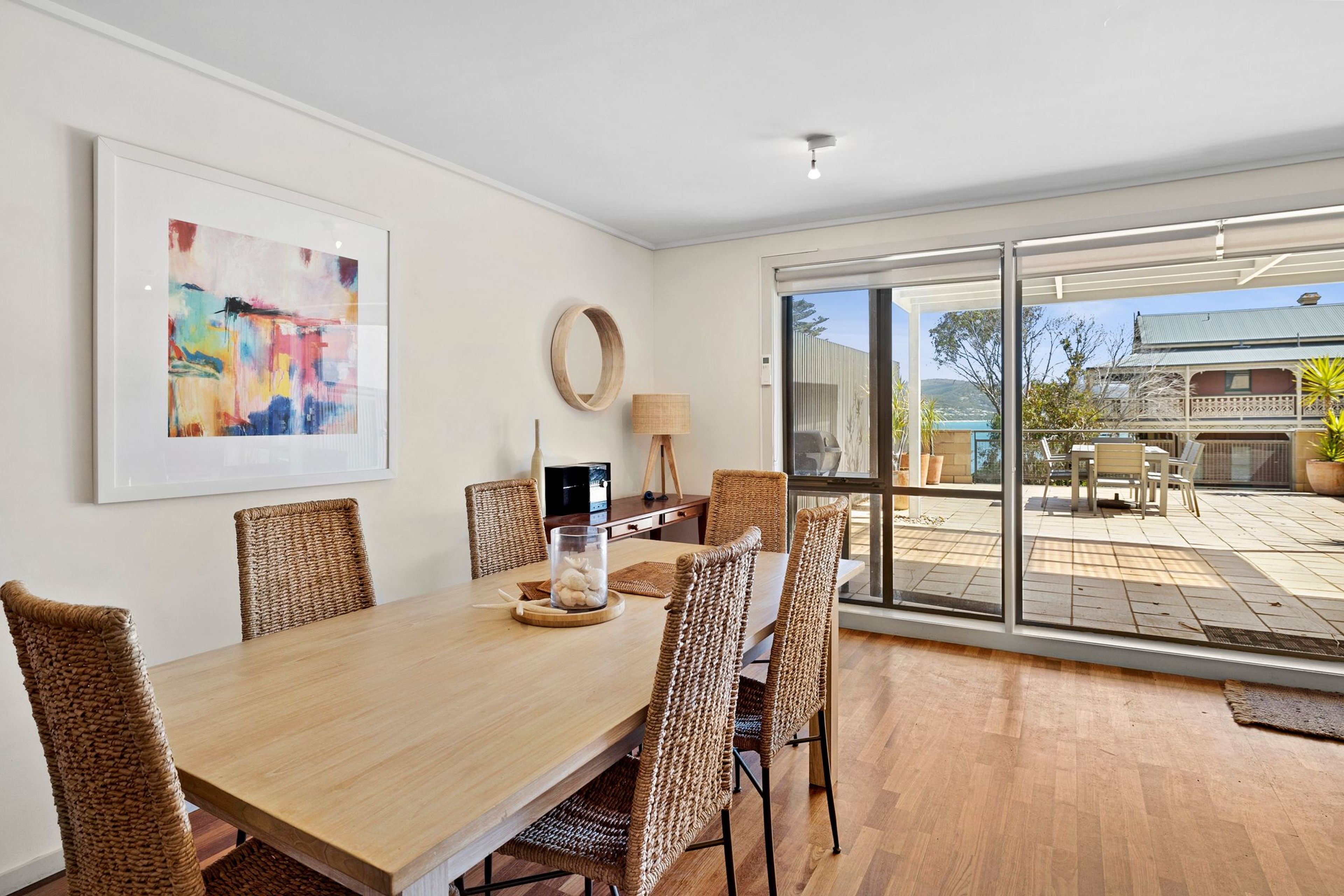 Un comedor con mesa de madera, sillas de mimbre, un cuadro abstracto enmarcado y vistas al exterior a través de grandes puertas correderas de vidrio.