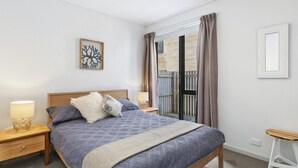 A bedroom with a bed, two bedside tables, a nightstand, a wall-mounted picture, and a window with curtains. - Apartment 23 Pacific Apartments (Lorne)