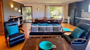 Ein Wohnzimmer mit einer Couch, Sesseln, einem Couchtisch und einem Essbereich mit Tisch und Stühlen. Es gibt eine Küchenzeile mit Schränken und einem Herd.