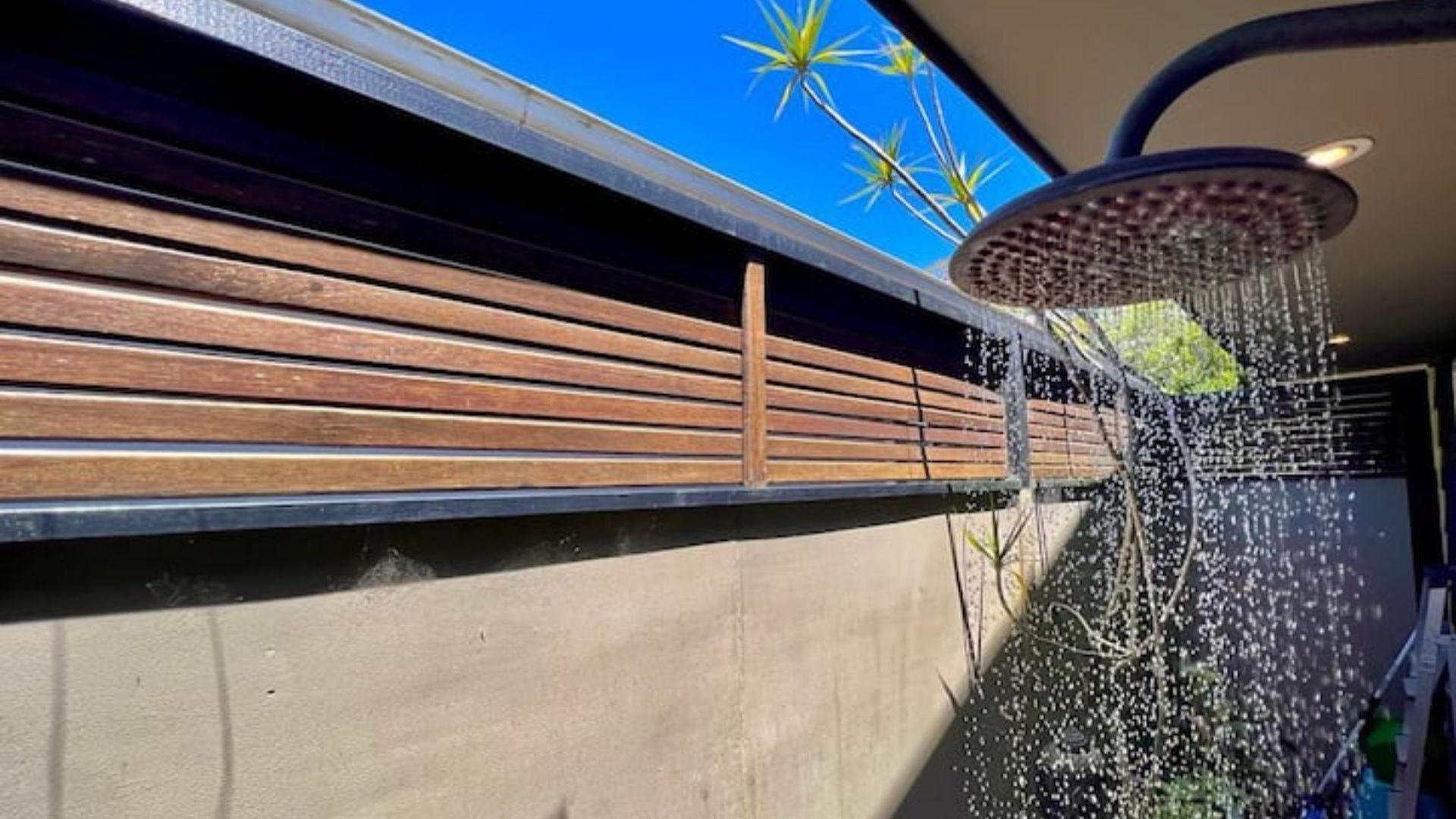 A shower with water spraying down, a wooden slatted wall, and a clear blue sky with palm trees in the background.