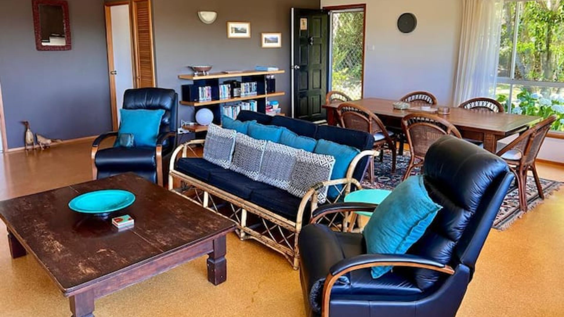 A cozy living room with a dark wooden coffee table, a blue sofa with patterned cushions, and a wicker chair. There is a bookshelf filled with books and decorative items, and a dining table with chairs.