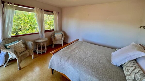 Une chambre à coucher avec un lit, deux fauteuils en osier, un petit tabouret et une fenêtre avec des rideaux.