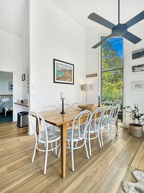 A dining area with a wooden table, white chairs, a ceiling fan, and a view of the outdoors. - SeaEsta - Modern Beachside Escape North Lorne (Lorne)