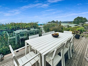 A white outdoor dining area with a table and chairs, surrounded by greenery and a glass railing.