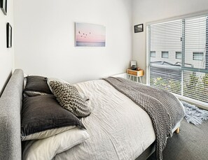 A bedroom with a bed, pillows, a bedside table, a lamp, a window with blinds, and a wall-mounted picture. - SeaEsta - Modern Beachside Escape North Lorne (Lorne)