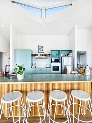 A modern kitchen with a central island, bar stools, and a view of the sky through skylights. - SeaEsta - Modern Beachside Escape North Lorne (Lorne)