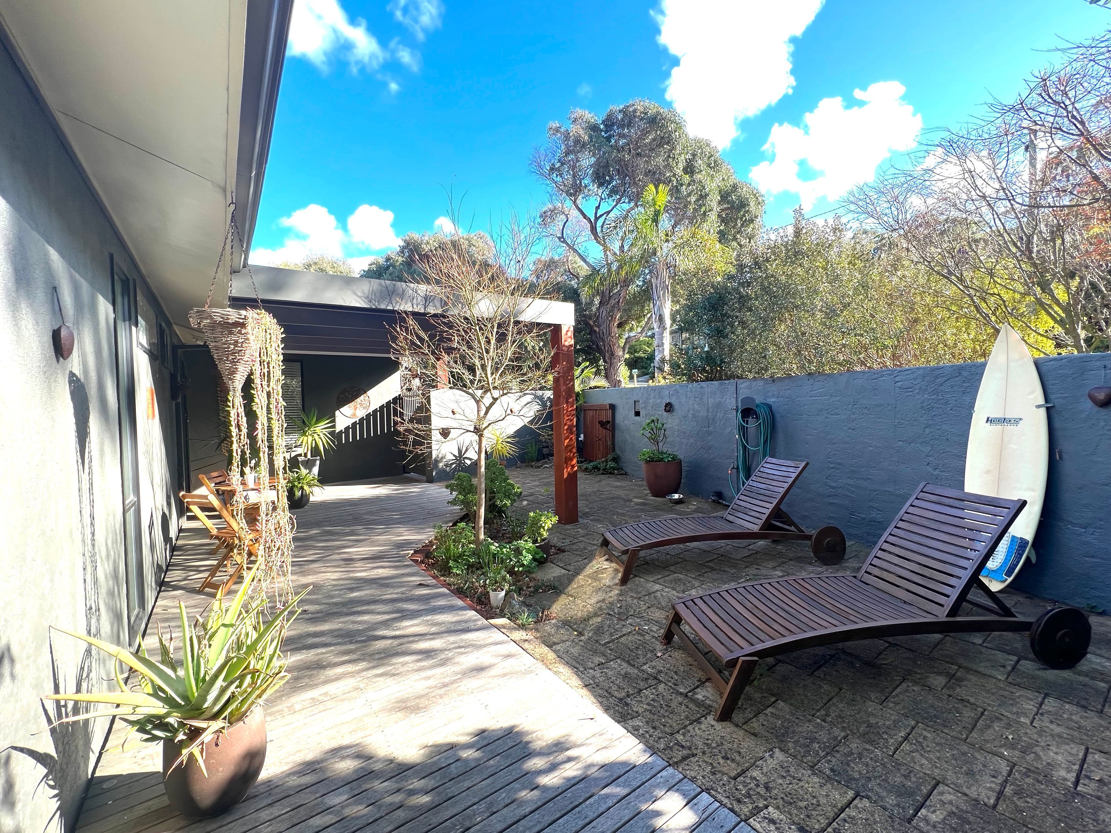 A sunlit outdoor area with a wooden deck, lounge chairs, potted plants, and a surfboard leaning against a wall.