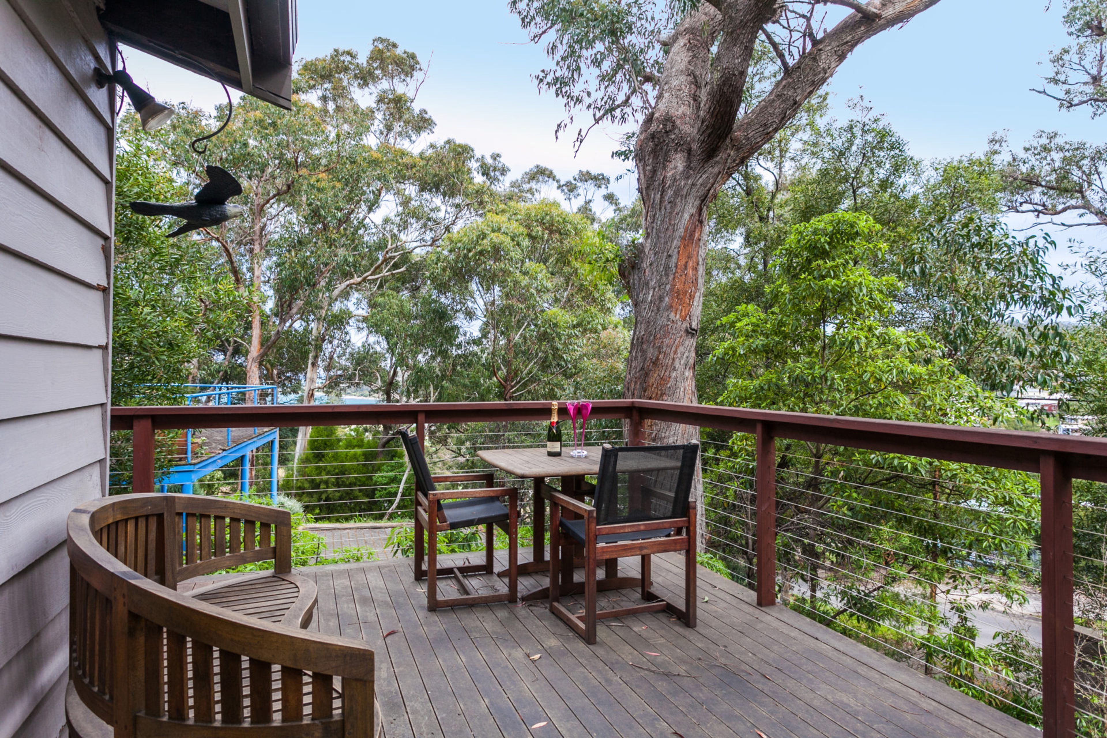 Terraza de madera con mesa y sillas, con vista a un paisaje verde y exuberante con árboles y una escalera azul.