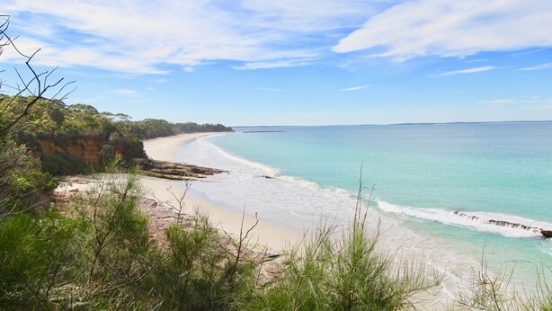 Un paisaje costero con una playa arenosa, aguas turquesas cristalinas y un acantilado rocoso con vegetación.