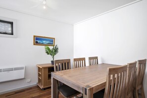 A dining area with a wooden table and chairs, a potted plant, a framed picture on the wall, and a window. - Apartment 36 Pacific Apartments (Lorne)