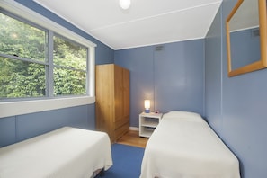A room with two beds, a wooden wardrobe, a nightstand with a lamp, and a window with a view of greenery.