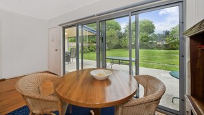Une pièce avec une table en bois et des chaises en osier, une porte-fenêtre coulissante donnant sur un espace extérieur avec une pelouse et une table.