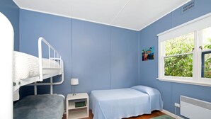 Une chambre avec un lit superposé, un lit simple, une table de chevet avec une lampe, un radiateur et une fenêtre donnant sur un espace verdoyant.