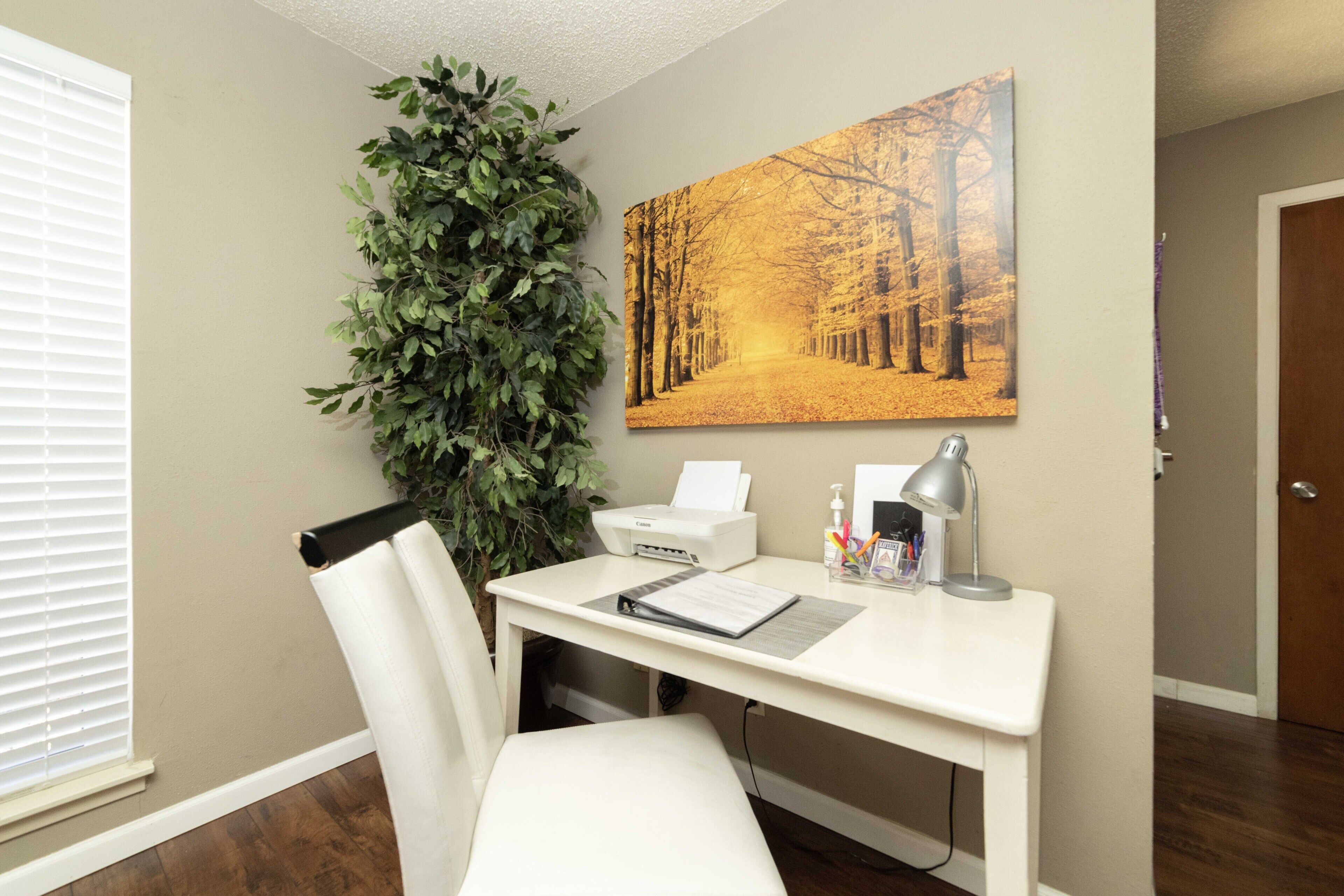 Ein Homeoffice mit Schreibtisch, Stuhl, Drucker, Lampe und einem großen gerahmten Bild von einem Wald.