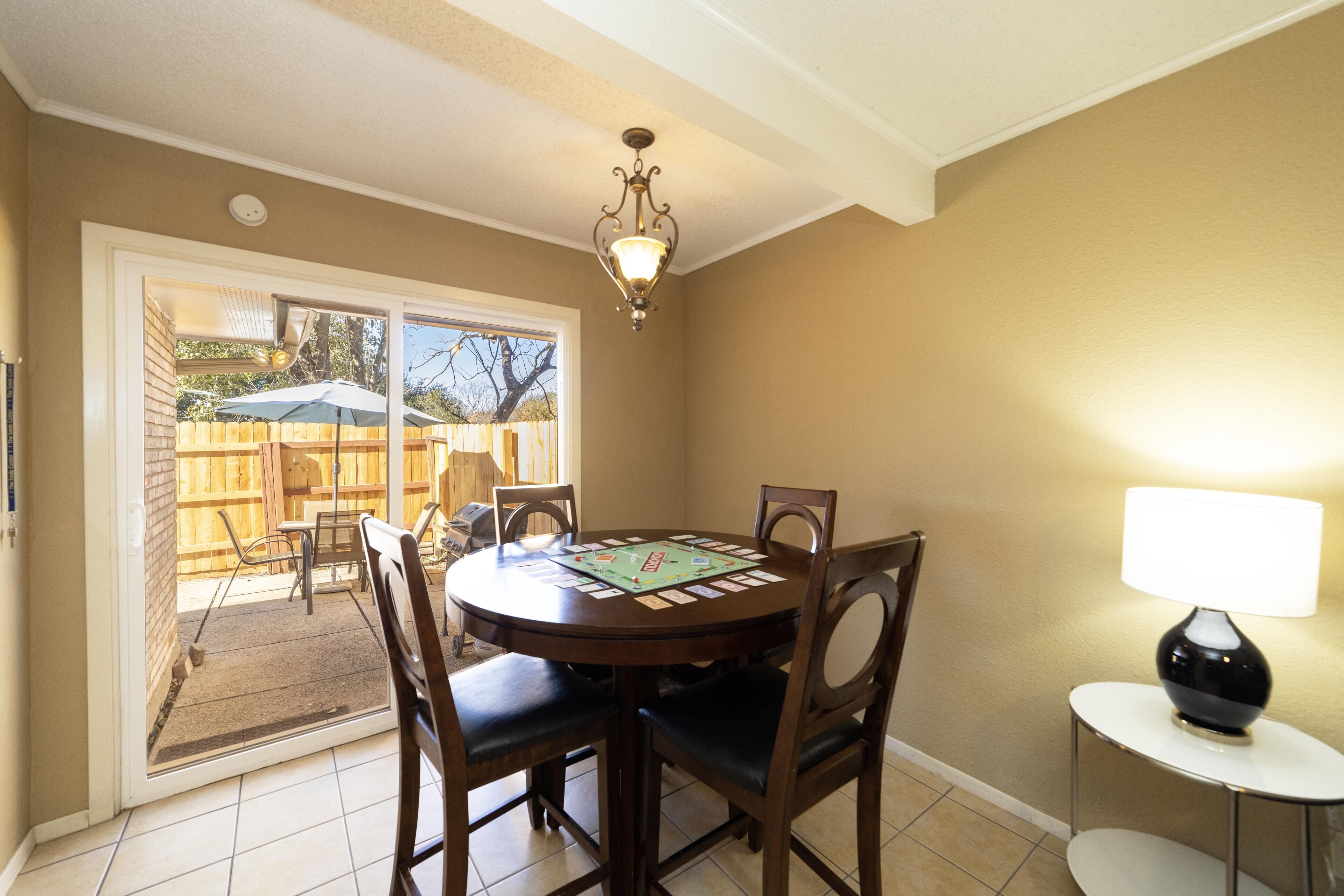 A dining area with a round table, chairs, and a chandelier. There is a sliding glass door leading to an outdoor patio with a wooden fence and chairs. A small table with a lamp is also visible.