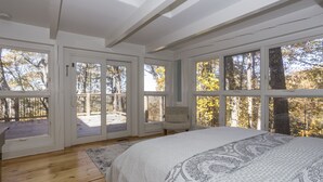 Un dormitorio con una cama grande, suelo de madera y varias ventanas que ofrecen vistas a árboles y una terraza.
