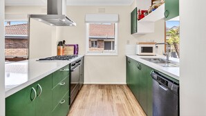 Une cuisine compacte équipée d’une cuisinière, d’un four à micro-ondes et d’un lave-vaisselle, avec des meubles de couleur verte et une fenêtre donnant sur un bâtiment en briques.