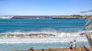 Une vue sur la côte, avec des vagues qui s’écrasent sur les rochers, une jetée qui s’avance dans l’eau et deux personnes qui se promènent le long de la plage.