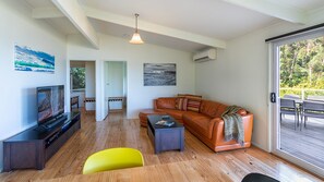 Ein modernes Wohnzimmer mit einer Ledersofa, einem Flachbildfernseher, Holzböden und einer Glasschiebetür, die auf eine Außenterrasse führt.