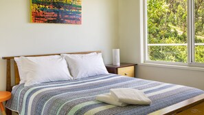 Ein Schlafzimmer mit Bett, Kissen, Nachttisch, Lampe und einem Fenster mit Blick auf Grünflächen.