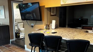 A kitchen with a TV mounted on the wall, a counter with bar stools, and a painting above the counter. - Beautiful 2BR 2BA Home with Private Pool & Jacuzzi - Includes Pool Table (Sarasota)