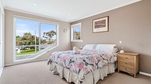 A bedroom with a large window, a bed with floral bedding, a nightstand, and a framed picture on the wall. - The Dunes 1 - Peaceful (Apollo Bay)