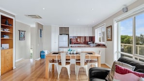 A modern kitchen with white cabinets, a dining area with a wooden table and white chairs, and a large window with a view of greenery. - The Dunes 1 - Peaceful (Apollo Bay)