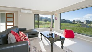 A modern living room with a black leather sofa, a glass coffee table, and a red bean bag. There is a large window offering a view of a green landscape.
