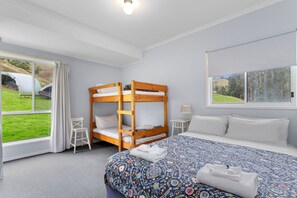 Ein Schlafzimmer mit Etagenbett, einem Einzelbett, einer gemusterten Tagesdecke, einem Fenster mit Blick auf Grünfl ächen und einem kleinen Tisch mit Lampe.