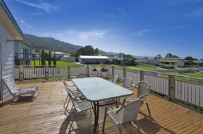 A wooden deck with a table and chairs, surrounded by a white fence, overlooking a residential area with houses and a mountain in the background. - Barrabay (Apollo Bay)