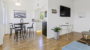 Una cocina moderna con armarios blancos, un comedor con una mesa y sillas redondas, y una sala de estar con un televisor montado en la pared.
