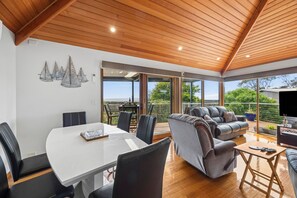 A modern living room with a dining area, wooden ceiling, and large windows offering a view of greenery.