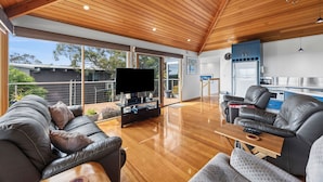 A spacious living area with a wooden ceiling, a flat-screen TV, leather sofas, and a kitchen area with a refrigerator and blue cabinets. - Blue Water (Anglesea)