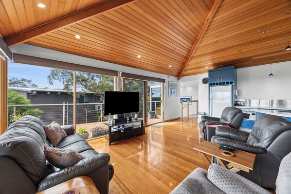 A spacious living area with a wooden ceiling, a flat-screen TV, leather sofas, and a kitchen area with a refrigerator and blue cabinets. - Blue Water (Anglesea)