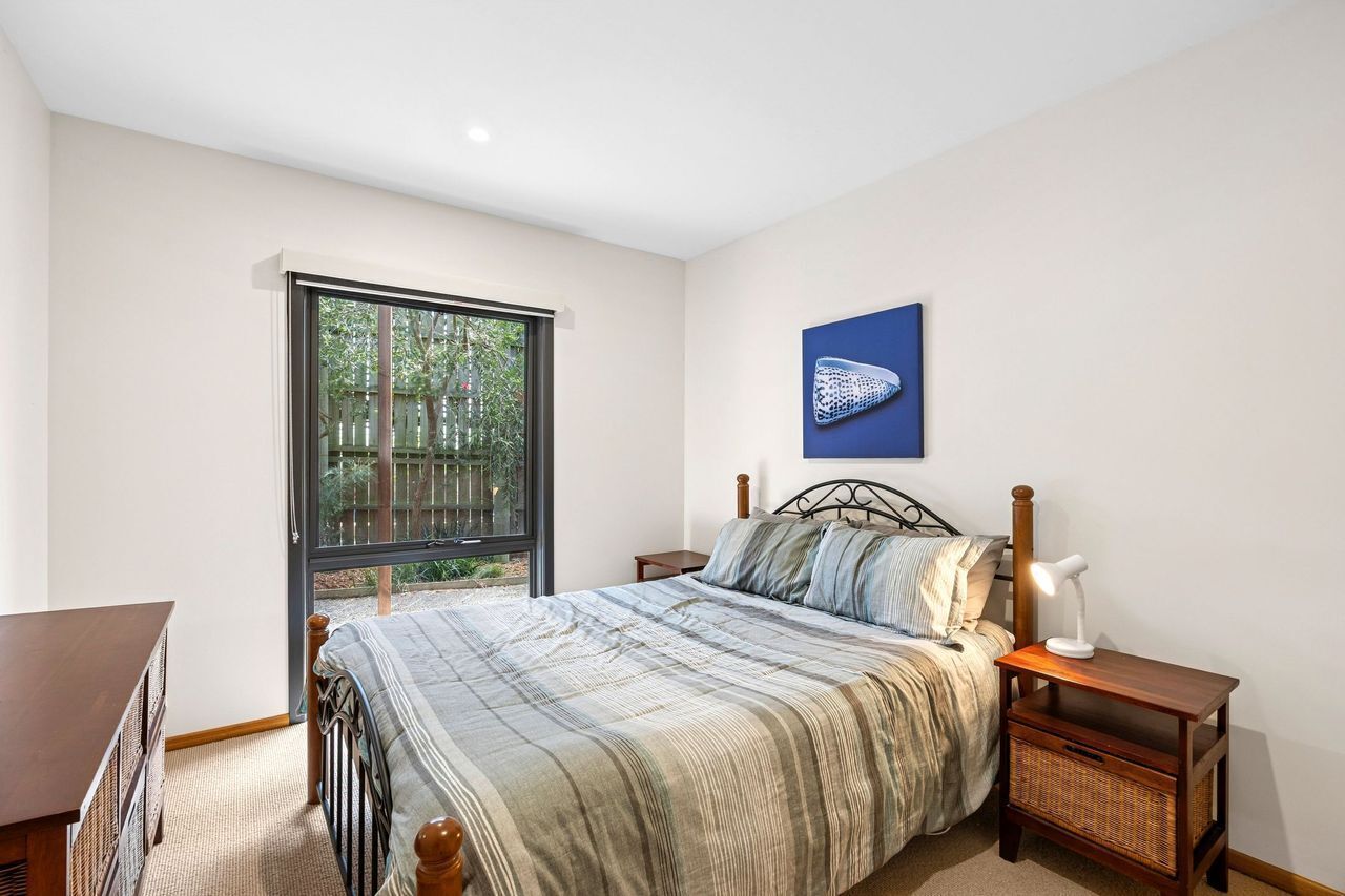A bedroom with a large window, a bed with a striped bedspread, a nightstand with a lamp, and a wall-mounted artwork.