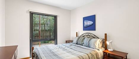 A bedroom with a large window, a bed with a striped bedspread, a nightstand with a lamp, and a wall-mounted artwork.