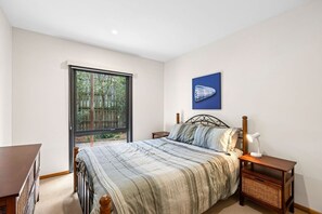 A bedroom with a large window, a bed with a striped bedspread, a nightstand with a lamp, and a wall-mounted artwork. - Blue Water (Anglesea)