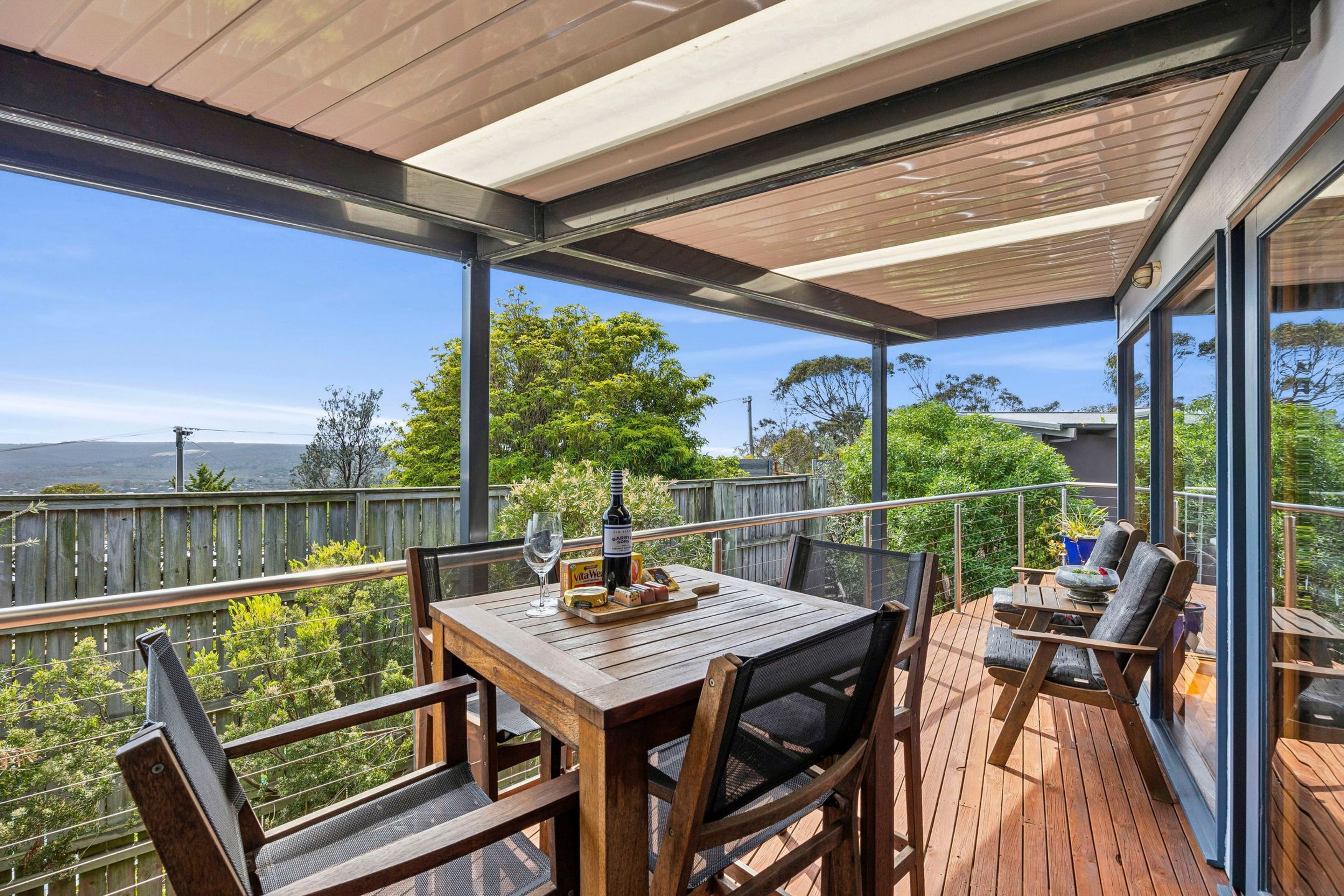 A covered outdoor patio with a wooden table and chairs, a bottle of wine, and a glass of wine.