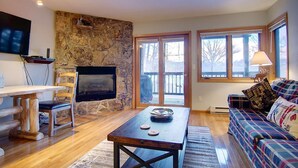 A cozy living room with a stone wall, a fireplace, a television, a wooden table, a sofa, and large windows. - Great Value! Epic Slope Views-Hot Tub+ Steps to Eat/Drink/Shops/Free Bus-W/D in Unit-Gas Fireplace (Steamboat Springs)